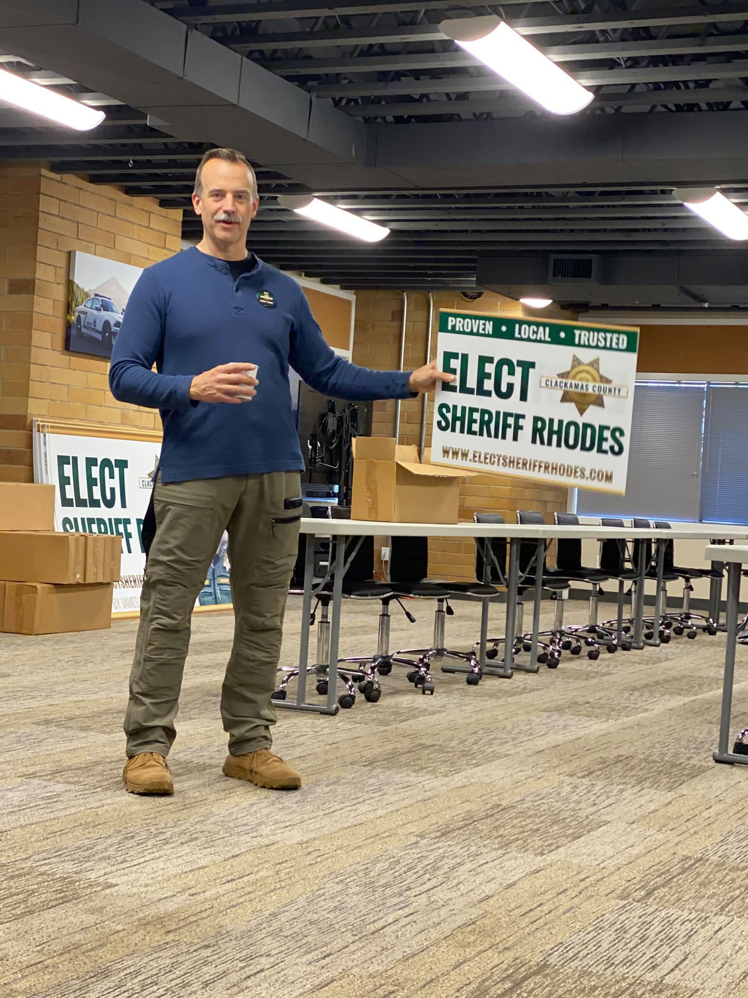 A man in a blue shirt holds an Elect Sheriff James Rhodes campaign sign in a meeting room.